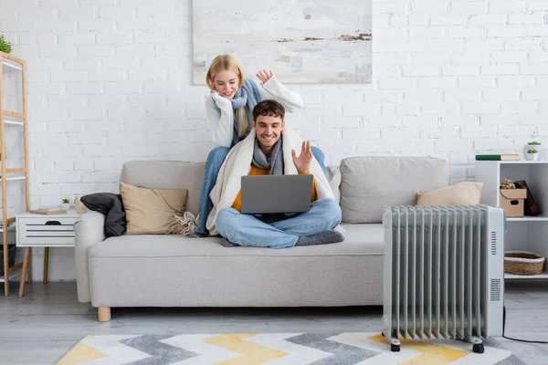 Heureux jeune couple en écharpes et pulls agitant les mains lors de l'appel vidéo sur ordinateur portable — Photo de stock