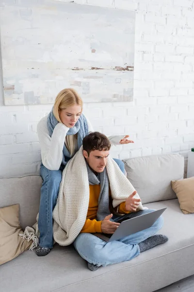 Displeased blonde woman in sweater and scarf sitting near freelancer and gesturing while looking at laptop — Stock Photo