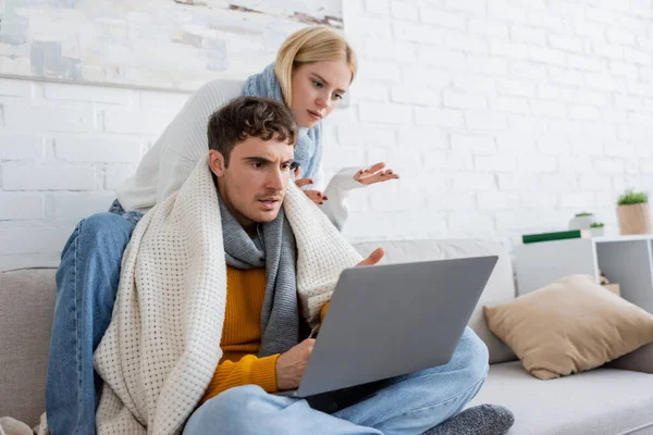 Mujer rubia disgustado en suéter y bufanda sentado cerca de novio y el gesto mientras mira el ordenador portátil - foto de stock
