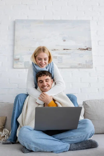 Mujer rubia feliz en suéter y bufanda abrazando novio mientras mira el ordenador portátil - foto de stock