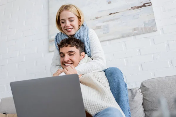 Mujer rubia feliz en suéter y bufanda abrazando novio alegre mientras mira el ordenador portátil - foto de stock
