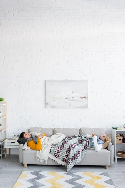 Jeune homme et femme blonde tenant des tasses et couché sous des couvertures sur le canapé dans le salon — Photo de stock