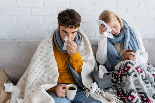 Sick couple covered in blankets holding cups of tea and tissues while sitting on sofa — Stock Photo