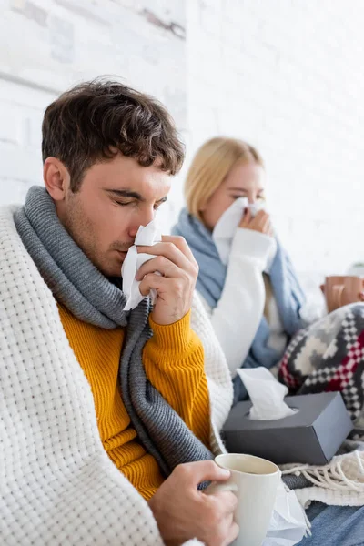 Sick man sneezing in napkin and holding cup of tea near blonde girlfriend in living room — Stock Photo