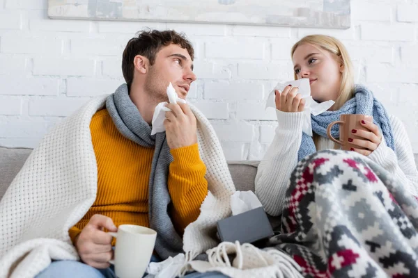 Diseased couple covered in blankets holding cups of tea and tissues while sitting on sofa — Stock Photo