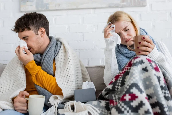 Erkrankter Mann niest neben blonder Frau mit Migräne und hält Tasse Tee — Stockfoto