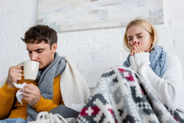 Kranke blonde Frau mit Schal niest in der Nähe eines Mannes, der Tee trinkt und Gewebe im Wohnzimmer hält — Stockfoto