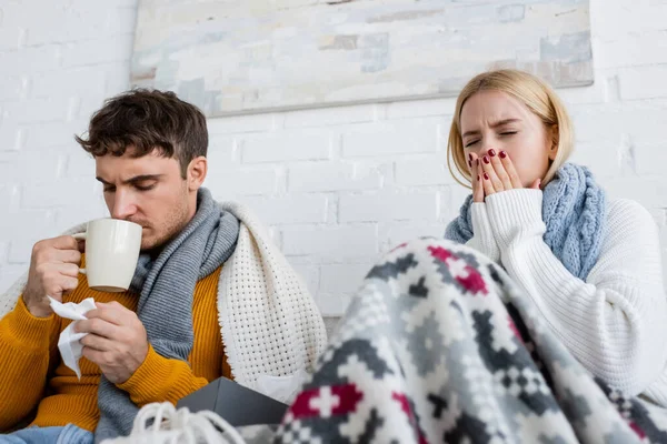 Femme blonde malade dans l'écharpe éternuer près de l'homme boire du thé et tenant des tissus dans le salon — Photo de stock