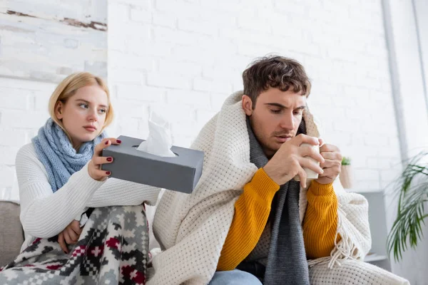 Blonde woman in scarf holding tissue box near sick boyfriend with cup of tea — Stock Photo