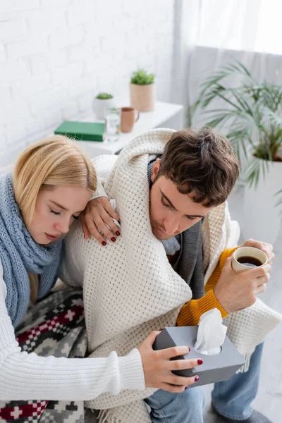 High angle view of sick blonde woman in scarf holding tissue box near diseased boyfriend with cup of tea — Stock Photo