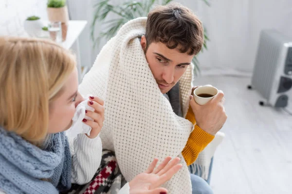 High angle view of sick blonde woman in scarf sneezing in tissue near diseased boyfriend with cup of tea — Stock Photo