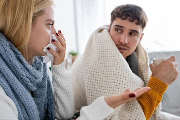 Sick blonde woman in scarf sneezing in tissue near diseased boyfriend with cup of tea — Stock Photo