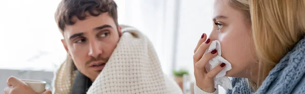 Sick blonde woman in scarf sneezing in tissue near diseased boyfriend with cup of tea, banner — Stock Photo