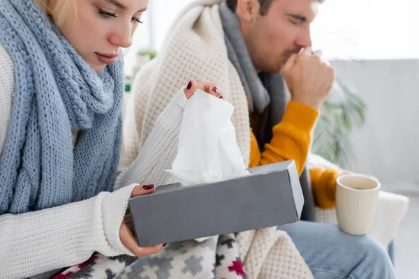 Kranke Frau nimmt Serviette aus Gewebebox in der Nähe verschwommener Mann hustet im Wohnzimmer — Stockfoto