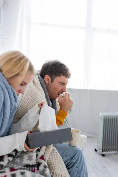 Homme malade avec les yeux fermés éternuer tout en tenant serviette et tasse près de petite amie — Photo de stock