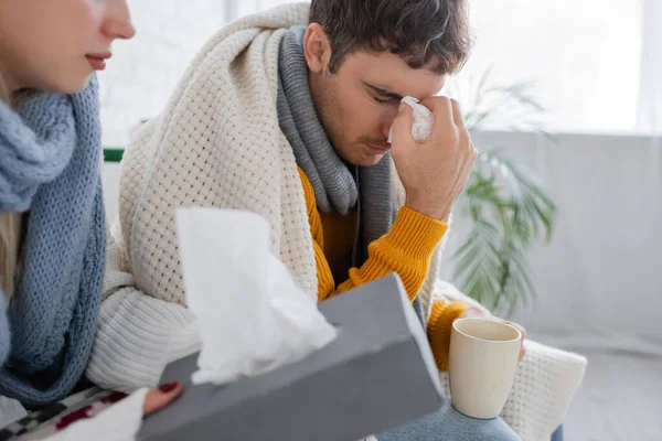 Kranker Mann mit geschlossenen Augen hält Serviette und Tasse in der Nähe Freundin mit Gewebebox — Stockfoto