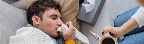 Top view of woman holding cup of tea near sick boyfriend lying near tissue box, banner — Stock Photo