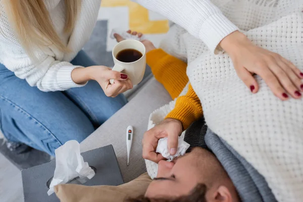 Vista aérea de la mujer sosteniendo taza de té cerca de novio enfermo acostado cerca de la caja de pañuelos - foto de stock