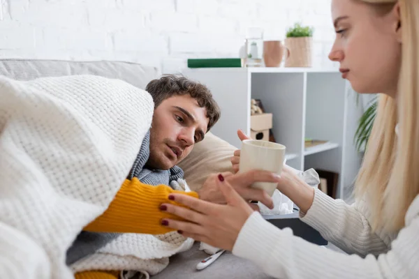 Donna premurosa che tiene una tazza di tè vicino al fidanzato malato sdraiato sul divano sotto la coperta — Foto stock