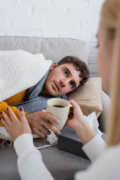 Novia cariñosa sosteniendo taza de té cerca de novio enfermo acostado en el sofá debajo de la manta - foto de stock