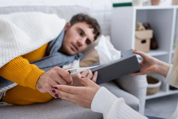 Cariñosa novia sosteniendo caja de tejido y termómetro digital cerca de novio enfermo acostado en el sofá debajo de la manta - foto de stock