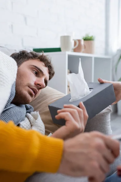 Petite amie attentionnée tenant boîte de tissu près du petit ami malade couché sur le canapé sous couverture — Photo de stock