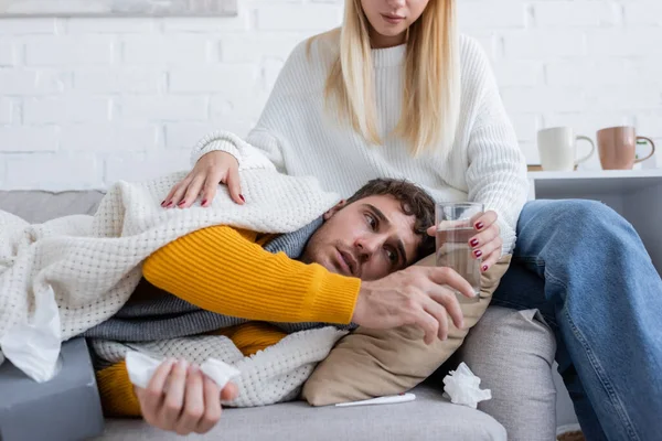 Blonde woman sitting on sofa and giving glass of water to sick boyfriend — Stock Photo