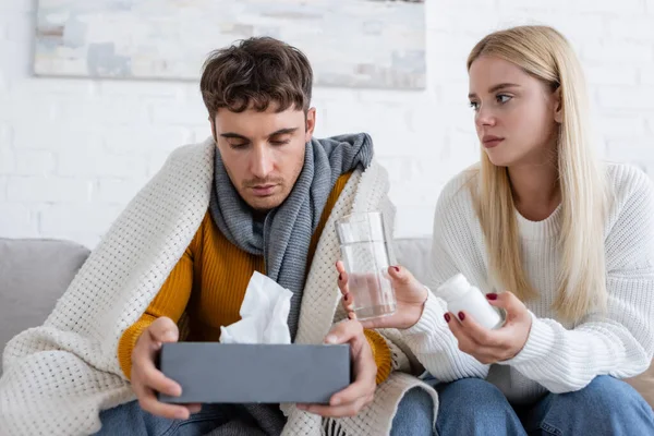 Cuidado joven mujer dando vaso de agua y botella con pastillas para novio enfermo con caja de pañuelos — Stock Photo