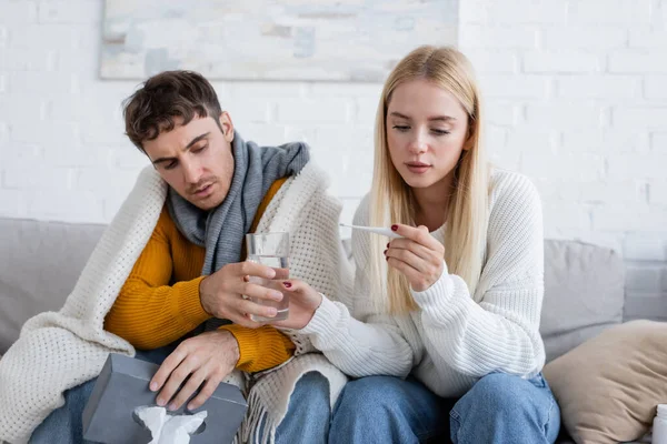 Young woman looking at digital thermometer and giving glass of water to sick boyfriend with tissue box — Stock Photo