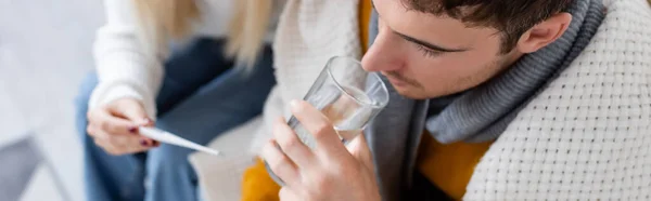 Vista superior del hombre enfermo beber agua cerca de la mujer sosteniendo termómetro digital, pancarta - foto de stock