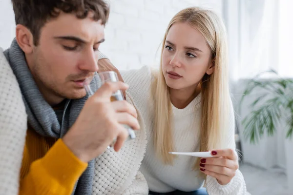 Hombre enfermo beber agua cerca de mujer preocupada sosteniendo termómetro digital - foto de stock