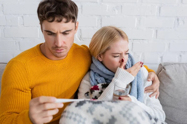 Mann hält digitales Thermometer in der Hand und umarmt kranke blonde Frau mit Glas Wasser — Stockfoto