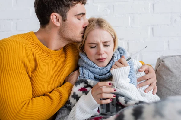 Homme étreignant et embrassant la tête de femme blonde malade avec verre d'eau et thermomètre numérique — Photo de stock