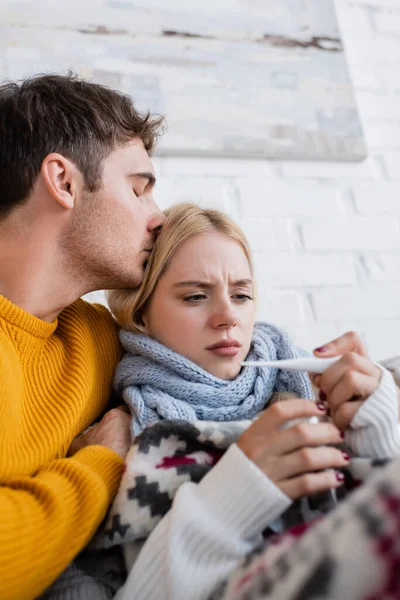 Uomo premuroso abbracciare e baciare la testa della donna bionda malata con termometro digitale — Foto stock