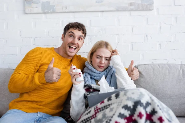 Homme joyeux montrant pouce près malade femme blonde avec boîte de tissu assis sur le canapé — Photo de stock