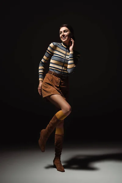 Full length of cheerful young woman in turtleneck with skirt and boots posing on black — Stock Photo