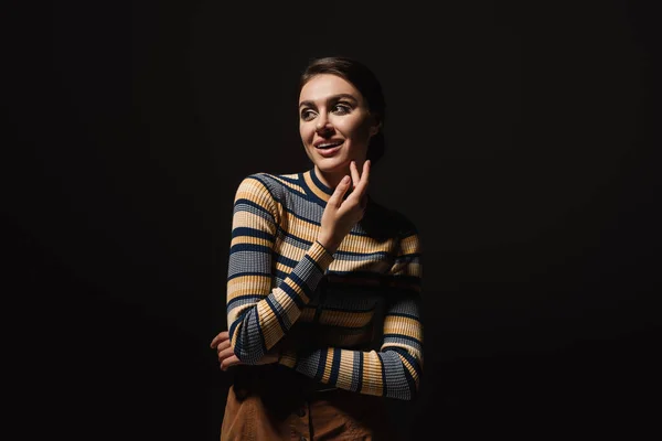 Alegre joven mujer en rayas cuello alto y falda mirando lejos aislado en negro — Stock Photo