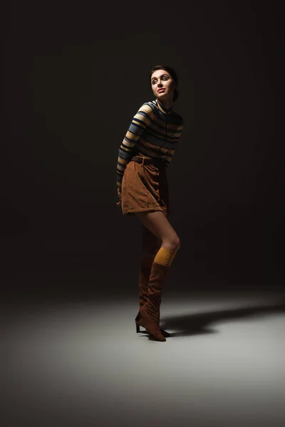 Full length of young woman in turtleneck with skirt and boots posing on black — Stock Photo