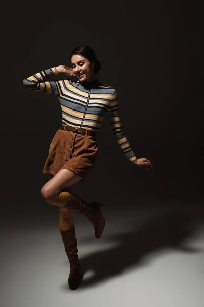 Pleine longueur de jeune femme heureuse à col roulé rayé avec jupe et bottes posant sur noir — Photo de stock