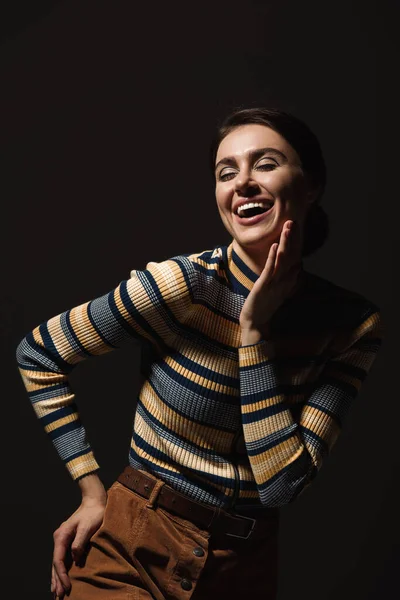 Pleased young woman in striped turtleneck and skirt posing with hand on hip isolated on black — Stock Photo