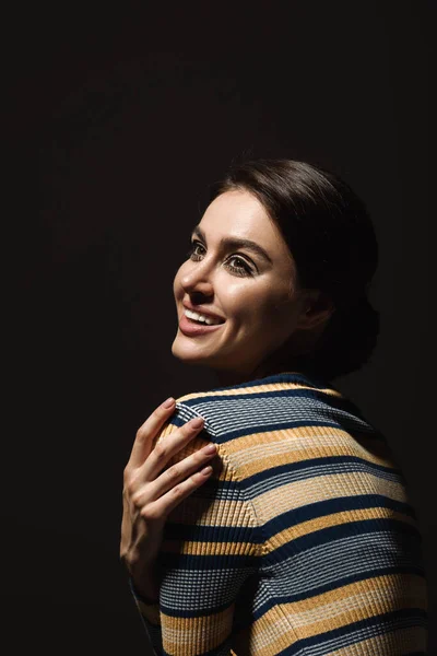 Positive jeune femme au col roulé rayé souriant et regardant loin isolé sur noir — Photo de stock