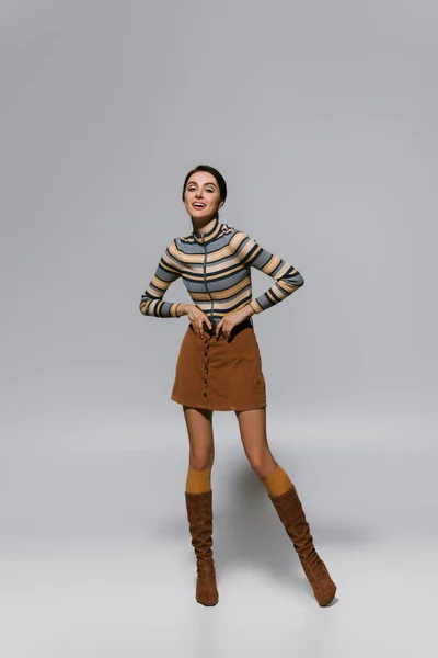 Full length of amazed young woman in striped turtleneck with skirt and boots posing on grey — Stock Photo