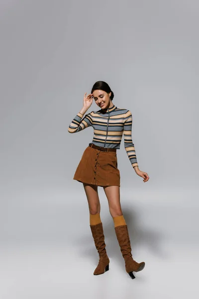 Full length of cheerful young woman in striped turtleneck with skirt and boots standing on grey — Stock Photo