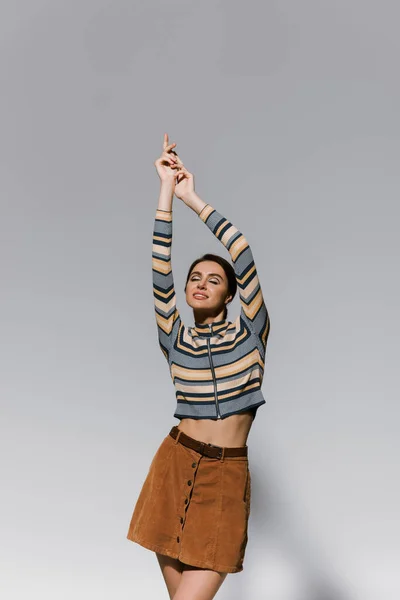 Happy young woman in striped turtleneck and skirt posing with raised hands isolated on grey — Stock Photo