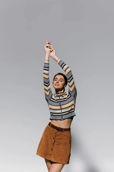 Heureuse jeune femme à col roulé rayé et jupe posant avec les mains levées isolé sur gris — Photo de stock