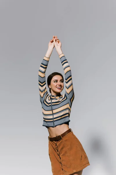 Fröhliche junge Frau in gestreiftem Rollkragen und Rock posiert mit erhobenen Händen isoliert auf grau — Stockfoto