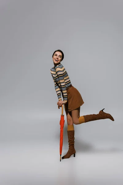 Full length of happy retro woman in autumnal outfit and boots posing with umbrella on grey — Stock Photo