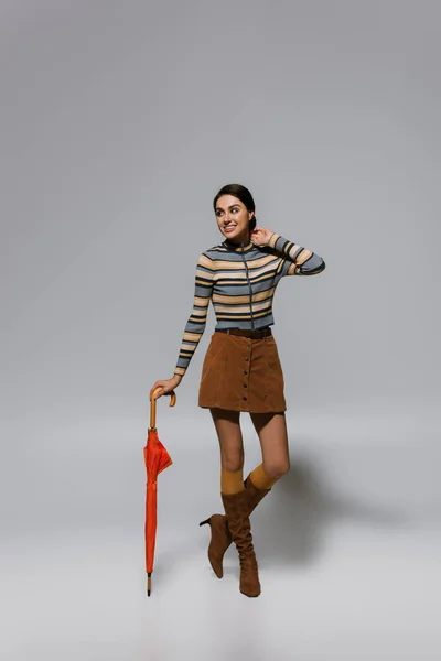 Full length of cheerful young woman in autumnal outfit and boots posing with umbrella on grey — Stock Photo