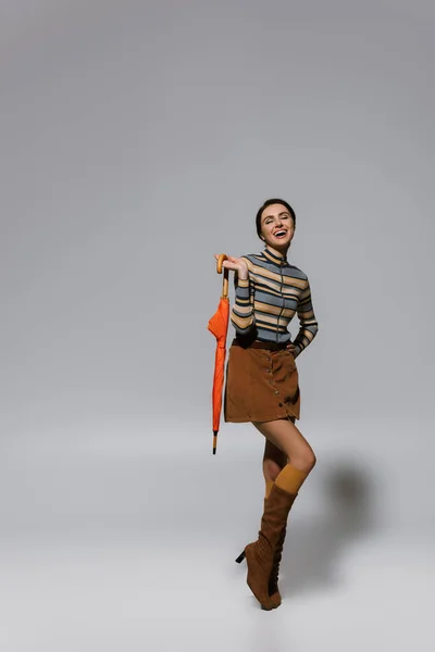 Повна довжина збудженої молодої жінки в автономному вбранні і чоботи позують з парасолькою на сірому — стокове фото