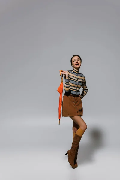 Longitud completa de la joven excitada en traje otoñal y botas posando con paraguas en gris - foto de stock